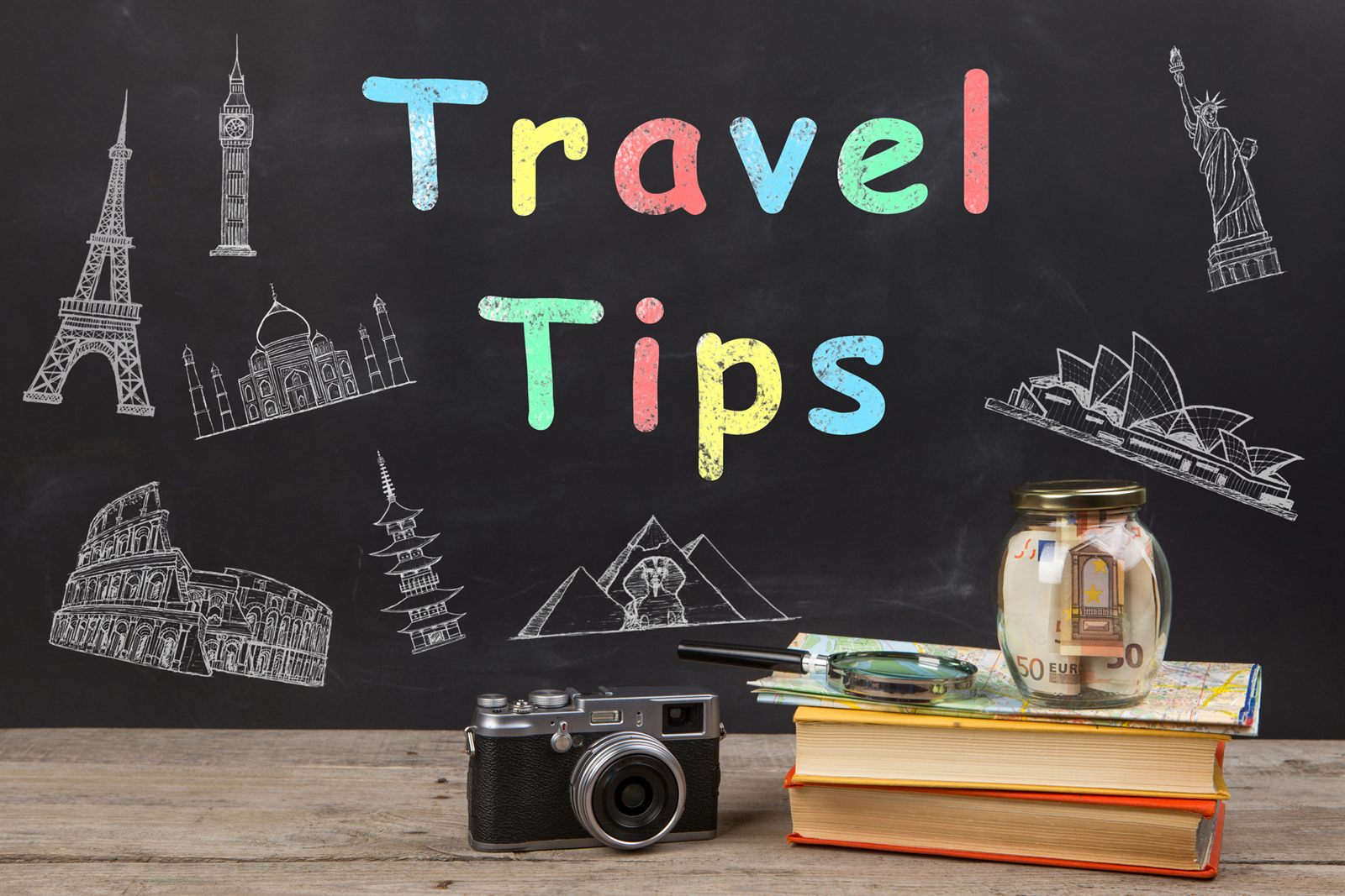 Chalkboard reading Travel Tips with a camera, books and a jar of saved banknotes